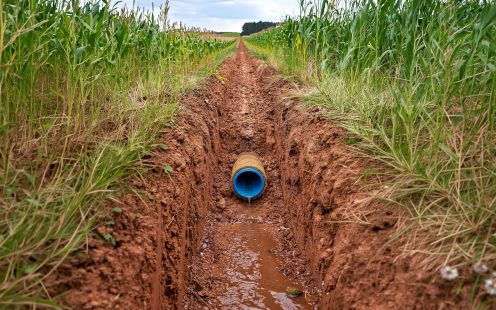 Zila gofrētā drenāžas caurule ar kokosšķiedru filtru iebūvēta lauksaimniecības tranšejā