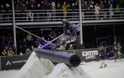 Frīstaila slēpotājs balansē uz melnas polietilēna caurules, kas novietota uz sniega tramplīna, fonā liels skatītāju pūlis.