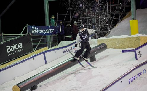 Frīstaila slēpotājs slīd pa lielu melnu polietilēna ūdensvada cauruli sacensību laikā un apkārt skatītāji.
