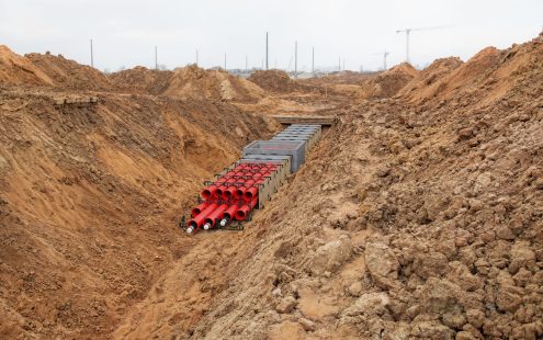 Dziļa būvbedre plašā būvlaukumā, kurā redzama OPI kabeļu kanālu bloku sistēmas izbūve. Tranšejas garumā stiepjas vairāki organizēti sarkanu kabeļu aizsargcauruļu saišķi atbalsta elementos. Tālāk tie ir savienoti ar dzelzsbetona vadu kanālu blokiem. Zemes kaudzes un būvkrāni tālumā liecina par apjomīgu infrastruktūras projektu.