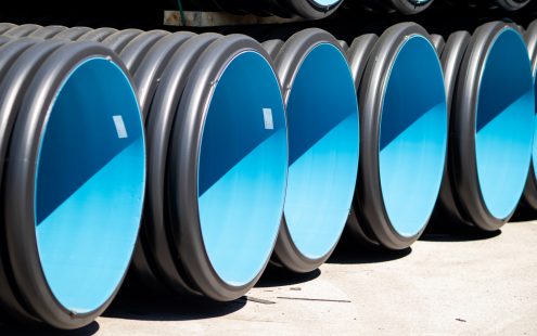 Tuvplānā rindā izkārtotas liela diametra melnas gofrētās caurules no 100% pārstrādāta PE, kurām ir gluda, gaiši zila iekšējā virsma.