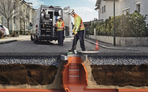 Šķērsgriezuma ilustrācija, kurā redzami divi strādnieki. Viens strādnieks no ielas ielaiž inspekcijas kameru Pipelife pazemes kanalizācijas akā, kamēr otrs strādnieks furgonā uzrauga procesu ekrānā. Šķērsgriezumā redzami grunts, grants un asfalta slāņi.